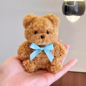 A small, handmade teddy bear plush named Tedd, crafted with curly, cinnamon-colored fabric and featuring a plaid bowtie. Its compact size fits in the palm of a hand, with a softly textured surface and a sweet, button-nosed expression.